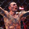 Max Holloway celebrating victory in the UFC octagon, arms raised, showing tattoos and wearing MMA gloves, with the cage and crowd in the background.