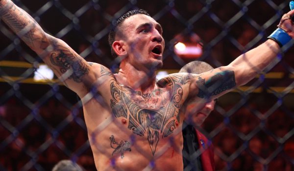 Max Holloway celebrating victory in the UFC octagon, arms raised, showing tattoos and wearing MMA gloves, with the cage and crowd in the background.